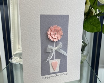 May include: A pink paper flower in a gray pot with a white ribbon and a pink heart. The card reads "happy mother's day".