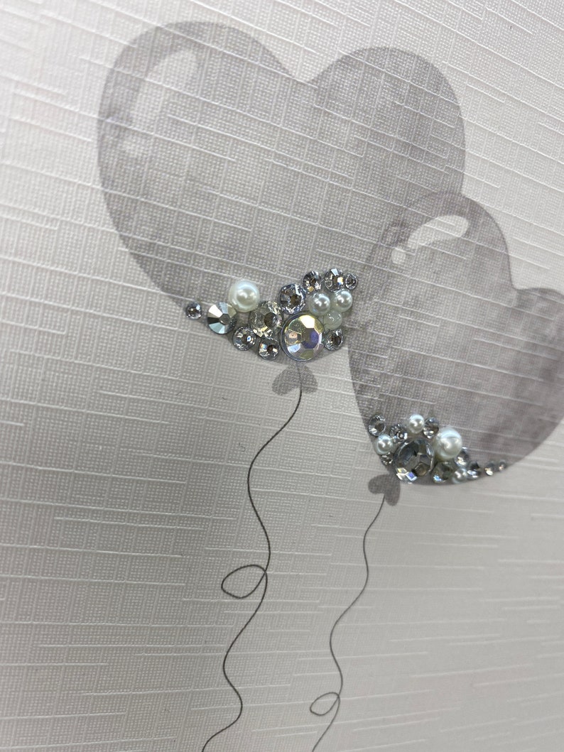 Pu&ograve; includere: Due palloncini grigi a forma di cuore con strass argentati e bianchi su uno sfondo bianco testurizzato.
