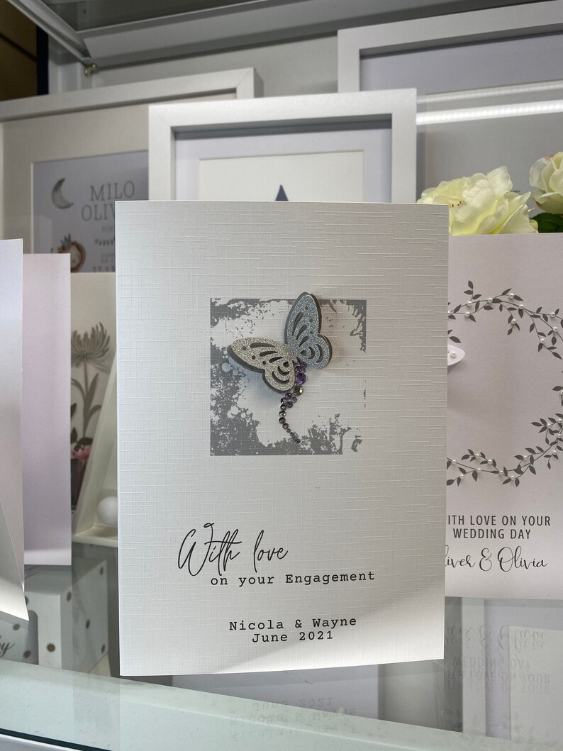 Pu&ograve; includere: Un biglietto bianco con un design a farfalla grigio e blu. Il biglietto recita "With love on your Engagement Nicola & Wayne June 2021".