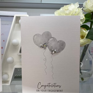 Pu&ograve; includere: Un biglietto bianco con uno sfondo testurizzato grigio. Due palloncini grigi a forma di cuore con dettagli di glitter argentati galleggiano sopra il testo "Congratulazioni per il vostro fidanzamento".