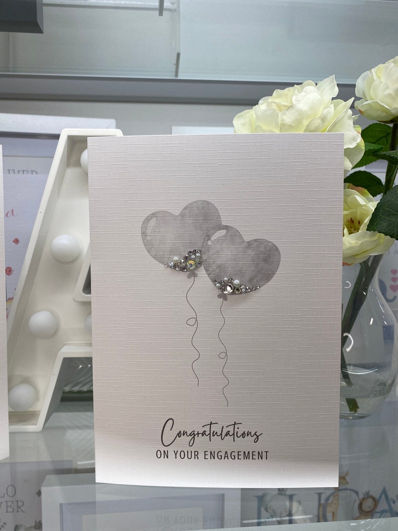 Pu&ograve; includere: Un biglietto bianco con uno sfondo testurizzato grigio. Due palloncini grigi a forma di cuore con dettagli di glitter argentati galleggiano sopra il testo "Congratulazioni per il vostro fidanzamento".
