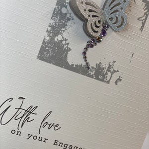 Pu&ograve; includere: Un biglietto bianco con una farfalla argentata con glitter e strass viola. Il biglietto dice "With love on your Engagement Nicola & Wayne"