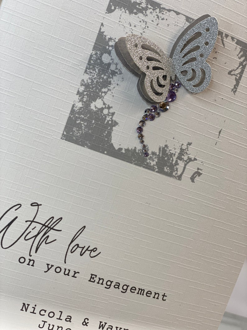 Pu&ograve; includere: Un biglietto bianco con una farfalla argentata con glitter e strass viola. Il biglietto dice "With love on your Engagement Nicola & Wayne"