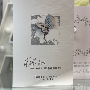 Pu&ograve; includere: Un biglietto bianco con una farfalla argentata glitterata su uno sfondo grigio. Il biglietto dice "With love on your Engagement Nicola & Wayne June 2021".
