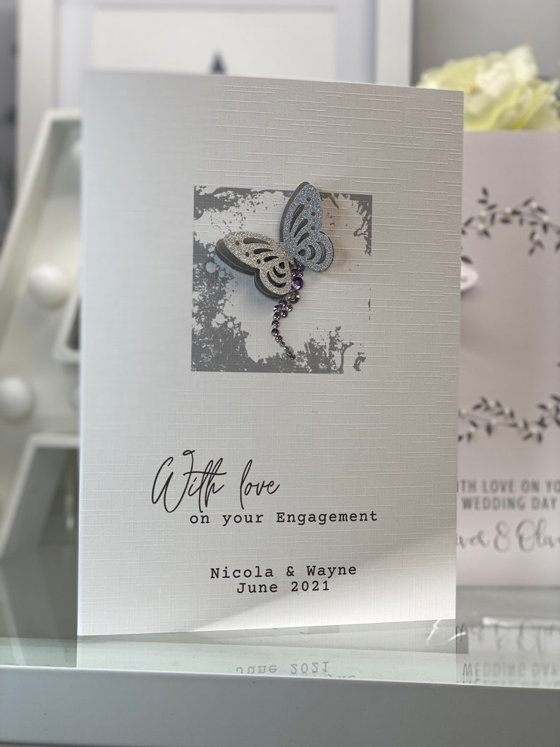 Pu&ograve; includere: Un biglietto bianco con una farfalla argentata glitterata su uno sfondo grigio. Il biglietto dice "With love on your Engagement Nicola & Wayne June 2021".