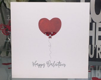 May include: A white card with a red heart-shaped balloon filled with red and white glitter. The card says "Happy Valentines" in black script.
