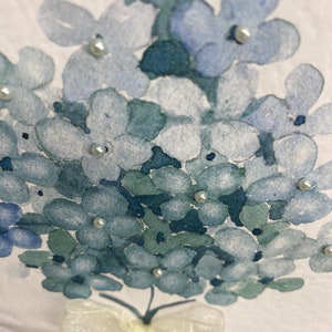 May include: A watercolor painting of a bouquet of blue hydrangeas with white pearl accents and a white satin bow.