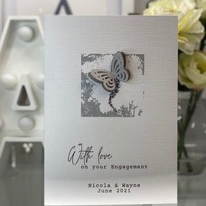 Pu&ograve; includere: Un biglietto bianco con una farfalla argentata glitterata su uno sfondo grigio. Il biglietto dice "With love on your Engagement Nicola & Wayne June 2021".