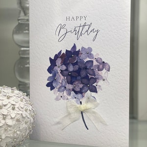 May include: A white card with the text "Happy Birthday" in cursive script. The card features a watercolor illustration of a bouquet of purple hydrangeas with a white satin ribbon.