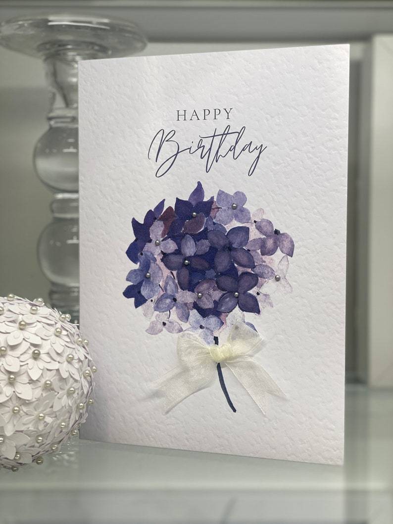 May include: A white card with the text "Happy Birthday" in cursive script. The card features a watercolor illustration of a bouquet of purple hydrangeas with a white satin ribbon.