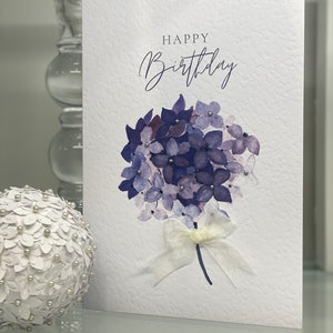 May include: A white card with the text "Happy Birthday" in cursive script. A watercolor painting of a bouquet of purple hydrangeas with a white ribbon is in the center of the card.