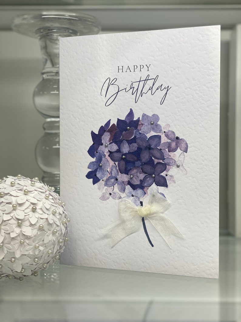 May include: A white card with the text "Happy Birthday" in cursive script. A watercolor painting of a bouquet of purple hydrangeas with a white ribbon is in the center of the card.