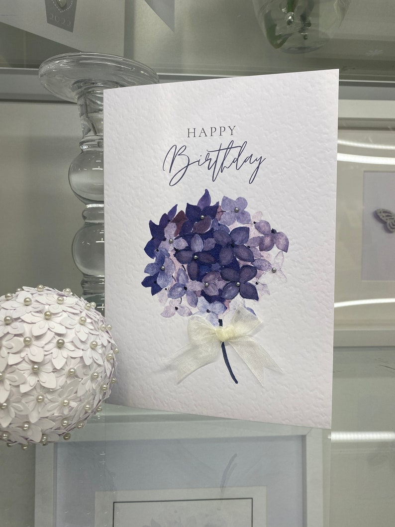 May include: A white card with the text "Happy Birthday" in black script. A watercolor painting of a purple hydrangea with a white ribbon is in the center of the card.