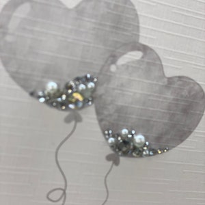 Pu&ograve; includere: Due palloncini grigi a forma di cuore con decorazioni argentate e bianche. I palloncini sono legati con dei nastri.