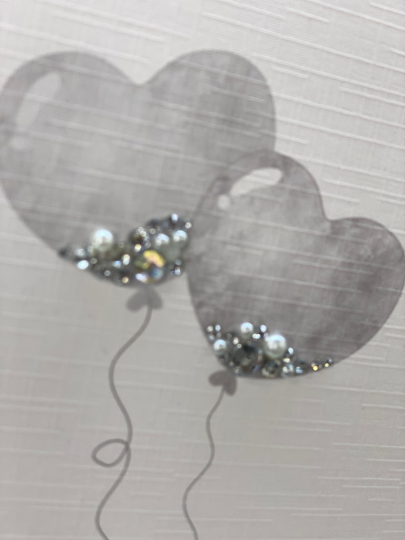 Pu&ograve; includere: Due palloncini grigi a forma di cuore con decorazioni argentate e bianche. I palloncini sono legati con dei nastri.
