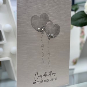Pu&ograve; includere: Un biglietto grigio con due palloncini grigi a forma di cuore con dettagli di glitter argentati. Il biglietto dice "Congratulazioni per il vostro fidanzamento".