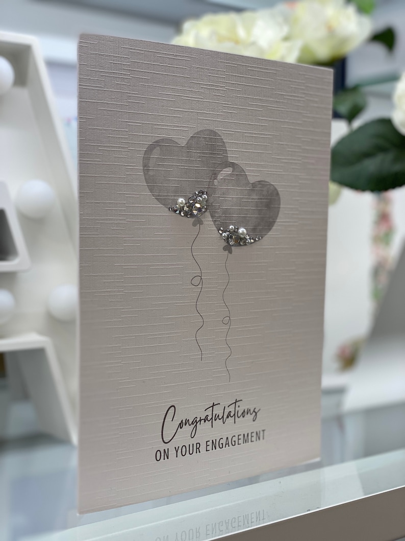 Pu&ograve; includere: Un biglietto grigio con due palloncini grigi a forma di cuore con dettagli di glitter argentati. Il biglietto dice "Congratulazioni per il vostro fidanzamento".