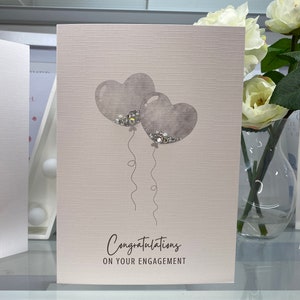 Pu&ograve; includere: Un biglietto bianco con una superficie testurizzata. Due palloncini grigi a forma di cuore con glitter argentati galleggiano. Il testo "Congratulazioni per il vostro fidanzamento" &egrave; stampato sulla carta.