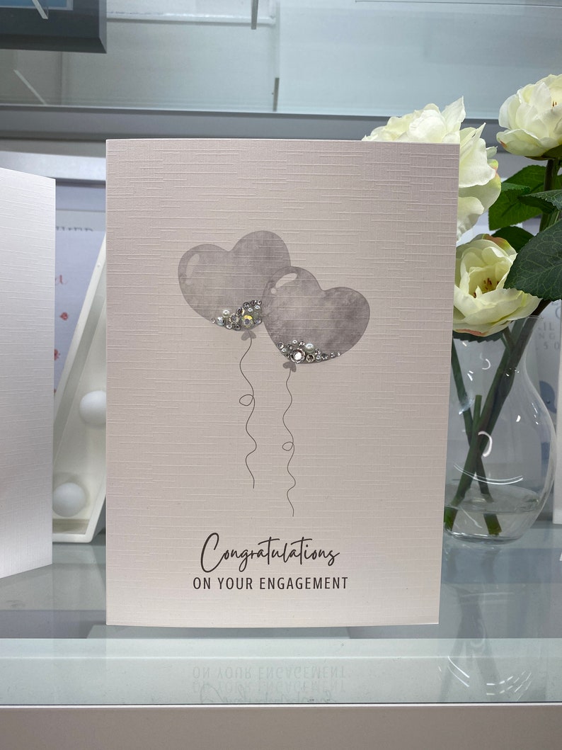 Pu&ograve; includere: Un biglietto bianco con una superficie testurizzata. Due palloncini grigi a forma di cuore con glitter argentati galleggiano. Il testo "Congratulazioni per il vostro fidanzamento" &egrave; stampato sulla carta.