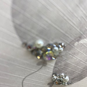 Pu&ograve; includere: Due grappoli di perline iridescenti e bianche, che ricordano palloncini, sono attaccati a delle stringhe su uno sfondo bianco testurizzato.
