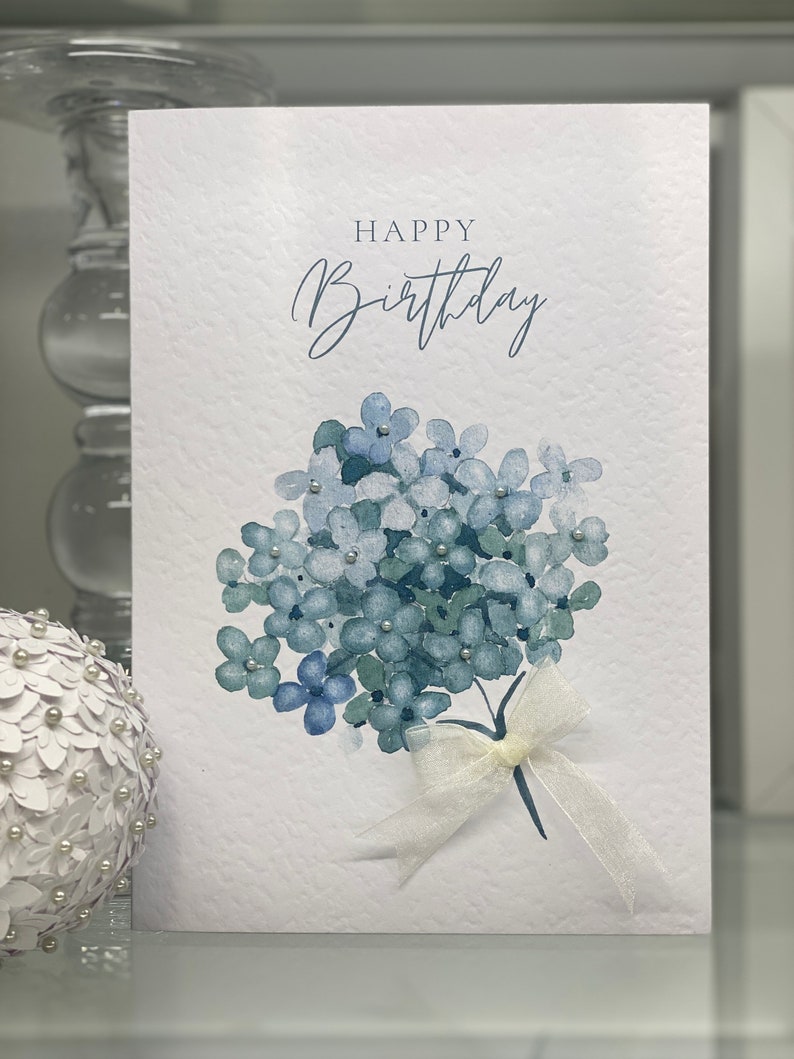 May include: A white card with the text "Happy Birthday" in blue script. A watercolor painting of a bouquet of blue flowers with a white ribbon is in the bottom right corner.