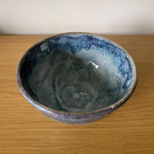 May include: A round ceramic bowl featuring a reactive glaze with deep blue and indigo tones at the rim that transition into a lighter, mottled grey-blue colour toward the centre. The bowl has a smooth, glossy finish and a simple, circular shape.