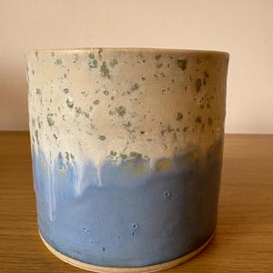 May include: A cylindrical ceramic mug with a two-tone glaze. The top half is a speckled off-white, transitioning to a light blue with dripped effects. The mug sits on a light-coloured wooden surface.