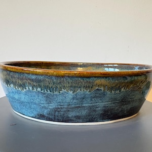 May include: A handmade ceramic bowl with an oval shape. The bowl has a glossy finish with a colour gradient, going from a deep blue at the base to a golden-brown rim. The bowl is approximately 30 cm long.