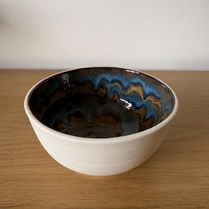 May include: A ceramic bowl with a matte off-white exterior featuring subtle horizontal ridges. The interior is finished with a glossy glaze in deep shades of blue, brown, and black, creating a flowing, layered pattern that drips down the sides.