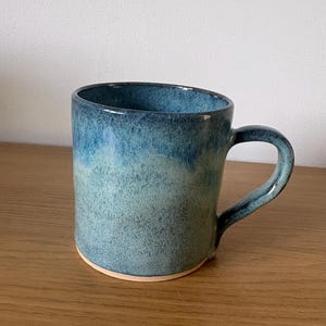 May include: A ceramic mug featuring a speckled glaze that transitions from a deep navy blue at the rim to a lighter, muted teal-green toward the base. The mug has a sturdy, curved handle and a visible unglazed clay rim at the bottom edge.