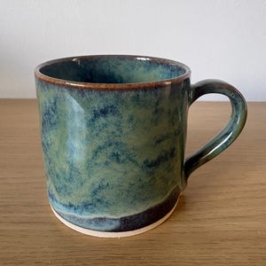 May include: A ceramic mug featuring a mottled glaze in shades of forest green, deep blue, and brown. The mug has a cylindrical body, a curved handle on the right side, and a smooth, unglazed light-coloured base that contrasts with the dark finish.