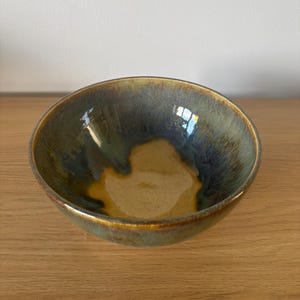 May include: A round ceramic bowl featuring a glossy glaze with blended tones of deep blue, forest green, and warm ochre. The interior surface displays a smooth, reflective finish with a distinct colour gradient transitioning from the dark rim to the centre.