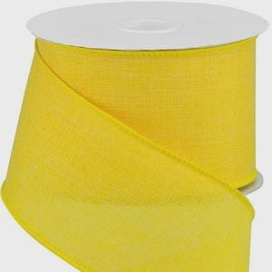 May include: A roll of bright yellow ribbon with a visible woven texture. The ribbon is wrapped around a white plastic spool, with a portion of the fabric cascading downward in a loose, elegant fold, showcasing the solid color and material.