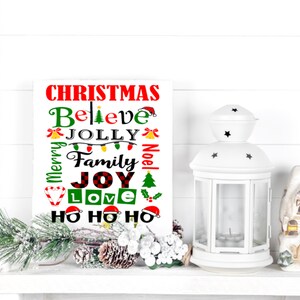 Puede incluir: Un letrero blanco con temática navideña con las palabras "Christmas, Believe, Jolly, Family, Joy, Love" en rojo, verde y negro. Una linterna blanca y una casa decorativa están a la derecha. Una rama de pino y piñas están a la izquierda.