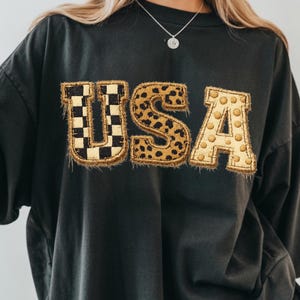 May include: Dark gray oversized t-shirt with the word "USA" in a decorative, textured font. The "U" features a black and white checkered pattern, the "S" has a leopard print, and the "A" has a gold polka dot design.