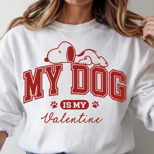 K&ouml;nnte beinhalten: Wei&szlig;es Sweatshirt mit roter Grafik, die Snoopy und den Text "MY DOG IS MY Valentine" zeigt. Das Design umfasst Pfotenabdr&uuml;cke und eine Schreibschrift. Ein l&auml;ssiges, bequemes Oberteil.