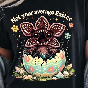 May include: Black t-shirt featuring a Demogorgon emerging from a cracked Easter egg. The egg is decorated with flowers and sits on grass. The text "Not your average Easter" is arched above the creature.