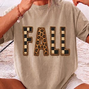 Könnte beinhalten: Ein beiges Kurzarm-T-Shirt mit dem Wort FALL in großen Blockbuchstaben auf der Brust. Die Buchstaben F, L und L haben ein schwarz-weißes Karomuster, während der Buchstabe A ein braunes Leopardenmuster aufweist.