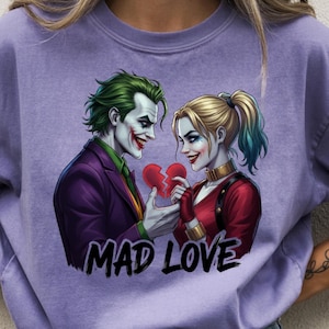Könnte beinhalten: Ein lilafarbenes T-Shirt mit einer Grafik von Joker und Harley Quinn, die ein zerbrochenes Herz halten. Der Joker trägt einen lila Anzug und grünes Haar, Harley Quinn ein rot-schwarzes Outfit. Der Text "MAD LOVE" steht unter dem Bild.