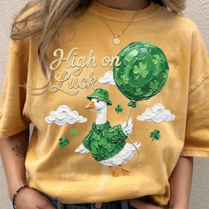 Puede incluir: Camiseta de color amarillo mostaza con la frase "High on Luck" en letras doradas. La camiseta presenta una ilustración caprichosa de un ganso con un suéter y sombrero verdes, sosteniendo un globo verde decorado con tréboles. Nubes blancas y tréboles completan el diseño.