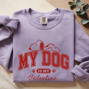 K&ouml;nnte beinhalten: Ein fliederfarbenes Sweatshirt mit einem roten Aufdruck von Snoopy und dem Text "MY DOG IS MY Valentine". Das Sweatshirt hat einen Rundhalsausschnitt und lange &Auml;rmel.