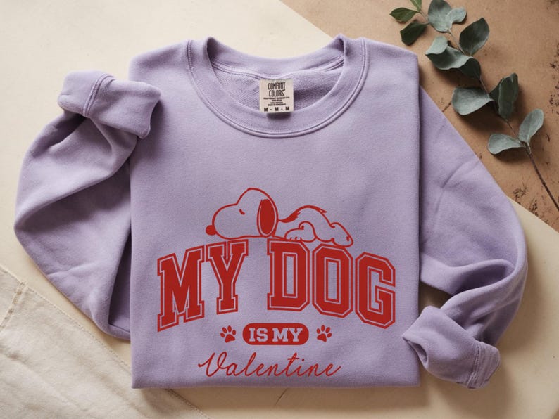 K&ouml;nnte beinhalten: Ein fliederfarbenes Sweatshirt mit einem roten Aufdruck von Snoopy und dem Text "MY DOG IS MY Valentine". Das Sweatshirt hat einen Rundhalsausschnitt und lange &Auml;rmel.