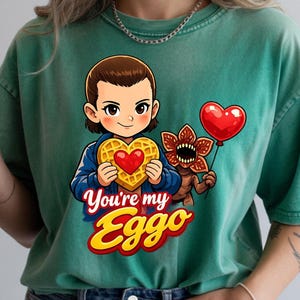 May include: Green t-shirt featuring a cartoon illustration of a character holding a waffle with a heart, a red heart balloon, and a Demogorgon. The text "You're my Eggo" is displayed in a colorful font.