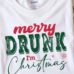 May include: White t-shirt with the words "merry DRUNK I'm Christmas" in red and green lettering. Two small snowflakes are printed on the shirt. The shirt is made of a soft material.