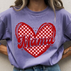 Könnte beinhalten: Ein lila T-Shirt mit einem roten Herzdesign und einem Schachbrettmuster. Das Wort "Mama" ist in einer Schreibschrift innerhalb des Herzens geschrieben. Das Shirt hat eine lockere Passform.