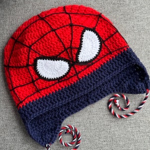 May include: Red and blue crocheted hat with a Spiderman design. The hat has a white crocheted eye mask and a black web design. The hat has two long ties.