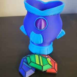 May include: A 3D-printed blue and purple decorative vase with a round purple accent and light blue accents. Several colorful, geometric, jewel-shaped objects in blue, green, and red are in front of the vase. The vase has a unique, futuristic design.