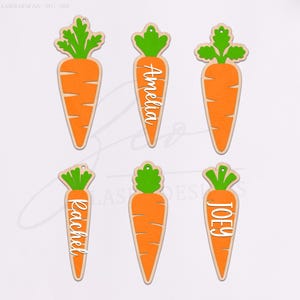 May include: Wooden carrot-shaped ornaments with green tops and orange bodies. Some carrots have names like "Amelia", "Rachel", and "Joey" in white script. The ornaments are likely for Easter or spring decorations.