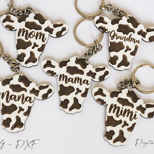 May include: Five wooden cow head keychains with brown and white cow print designs. Each keychain has a different name engraved on it: Mom, Grandma, Mama, Nana, and Mimi.