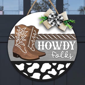 May include: Round wooden door sign with a country theme. Features brown cowboy boots, the words "HOWDY Folks", and a black and white cow print. A burlap and plaid bow with greenery is attached.